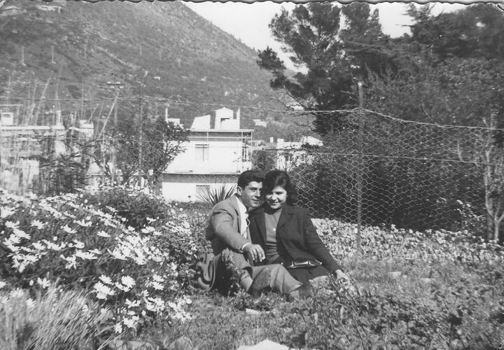 Man and woman smile as they sit outdoors amongst flowers. Mountainside behind them.