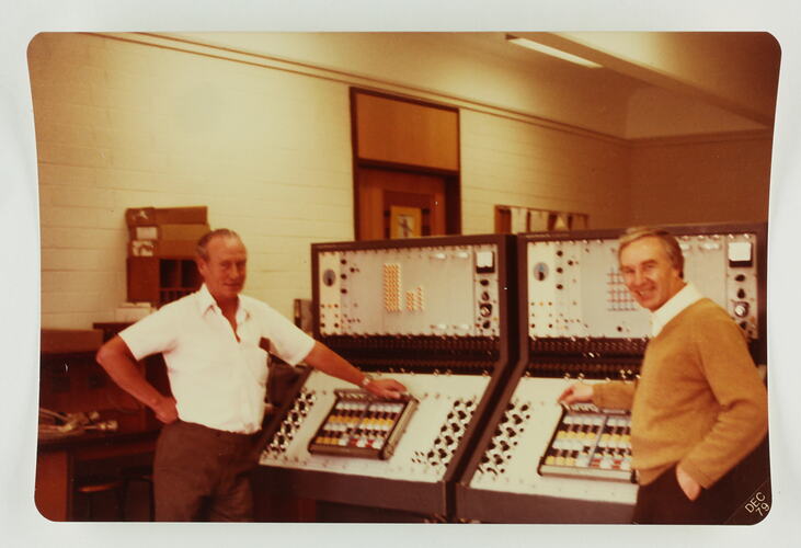 Two men pose in front of two large computers.