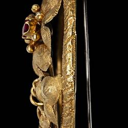 Profile of gold brooch with large flower with red centre, supported by two lower leaves and tendrils, inside an oval border. Vertical pin.