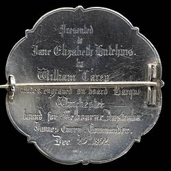 Back of brooch made from silver coin. Scrolled edge. Raised lettering. Pin across centre.