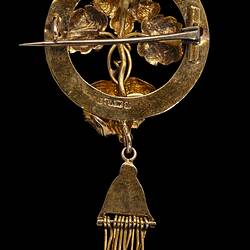 Back of round gold brooch. Horizontal central pin closed. Tassel hangs from base.
