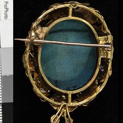Back of round gold brooch with gold border and horizontal pin. Back is green.