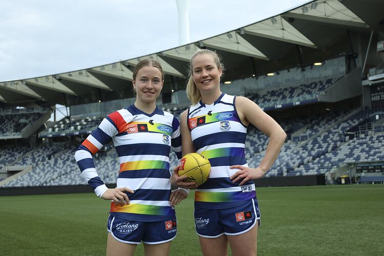 Two female footballers pose wearing Geelong guernseys. Grandstand behind.