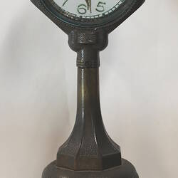 Clock with clear glass face on dark green painted stand.