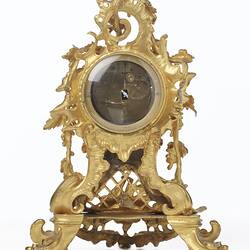 Back of elaborately decorated gold mantel clock. Round glass door shows internal mechanism.