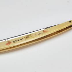 Cut throat razor, metal blade, ivory-styled plastic handle with 'PHAR LAP', picture of jockey on racehorse and two flags. Closed.
