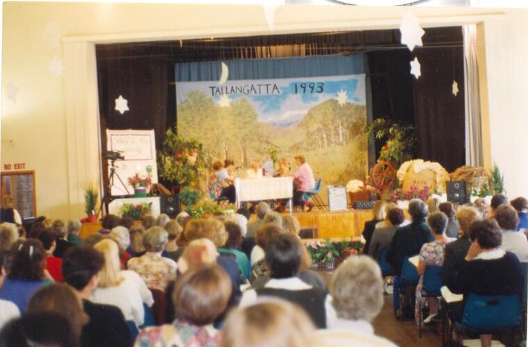 View from behind of seated crowd watching women seated at table on stage.