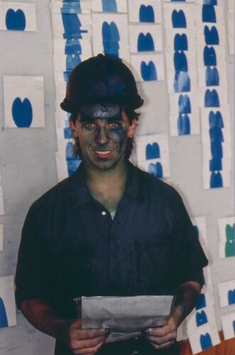 Man wearing blue boiler suit and safety helmet. Behind him is a chart with blue samples. He has blue paint on