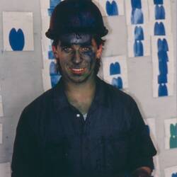 Man wearing blue boiler suit and safety helmet. Behind him is a chart with blue samples. He has blue paint on