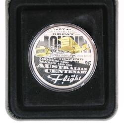Coin, round, yellow flying biplane. Text behind and below. Housed in black velvet case.