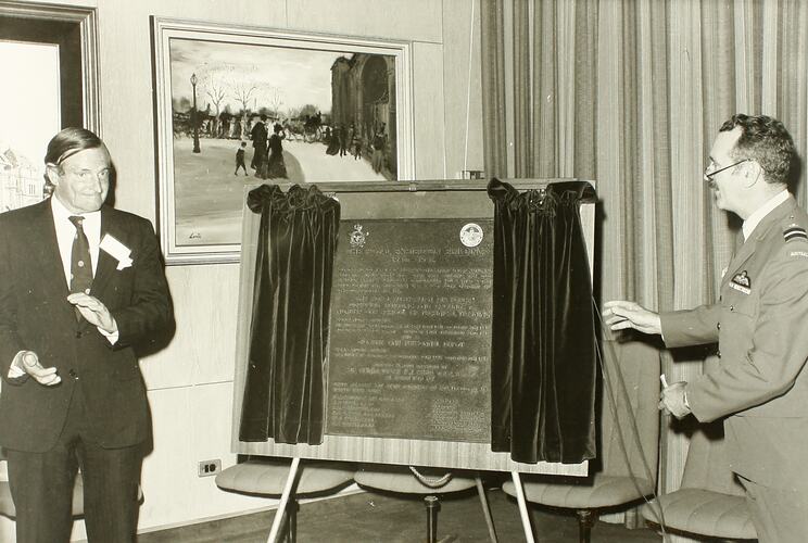 Black and white image of two men in suits unveiling commemorative plaque on an easel with velvet curtains.