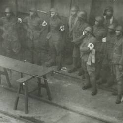 Group of uniformed men with helmets and red cross bands on their arms, stretcher on left.