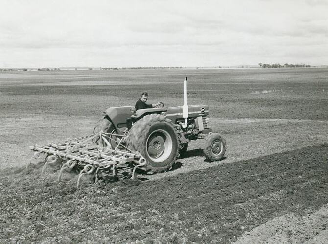 Side view of a man driving a tractor coupled to a cultivator in a large open field.