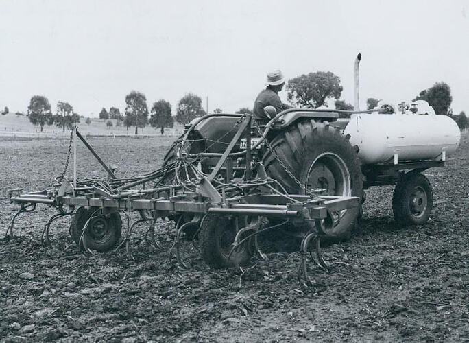 Rear view of a man driving a tractor fitted with a liquid fertilizer tank and a tine cultivator.