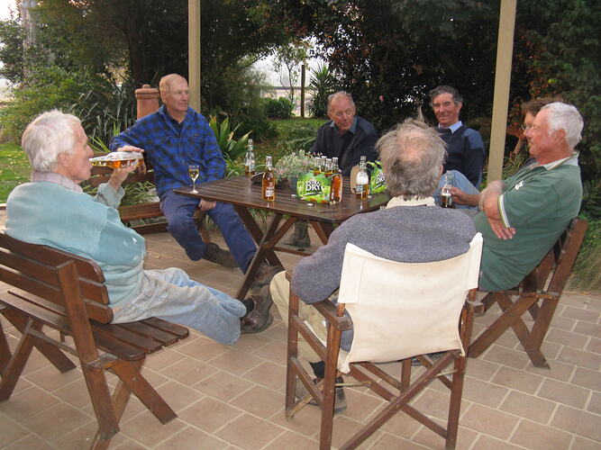 Seven older men sitting around table drinking beer.