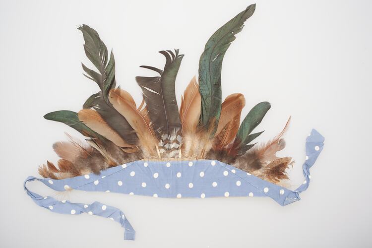 Feathered headdress. Brown and green feathers mounted in a blue and white spotted band with green bow.