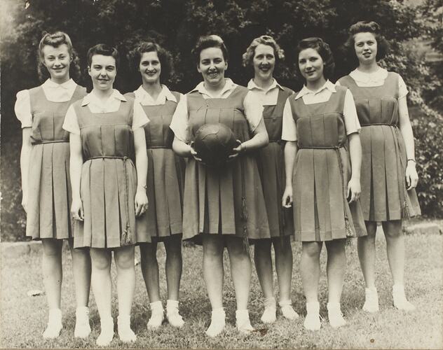 Seven women in basketball tunics standing in an outdoor setting. The middle woman holds a basketball.