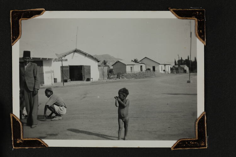 Naked boy standing in a street, with men to the left and row of houses in background.