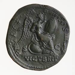 Roughly round coin. Seated winged figure facing right on a shield, she is inscribing on a second shield which rests on her lap. Text around.