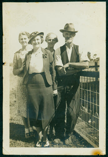 Man with a pipe in his mouth standing propped against a fence with three women to his right.