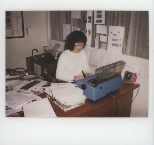 Woman at crowded desk, typing on typewriter.