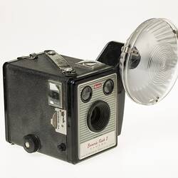 Box camera with black body. Oversized circular flash at top left corner.