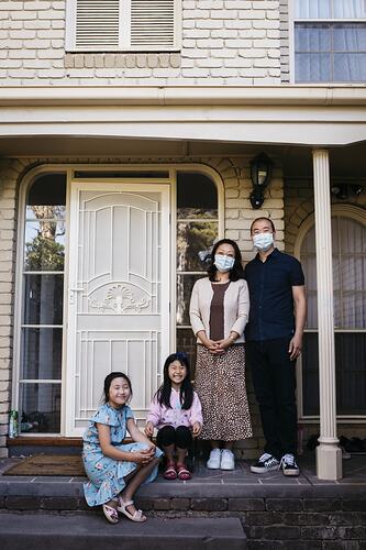 Woman, man and two girls pose on front porch of house. The woman and man wear masks.