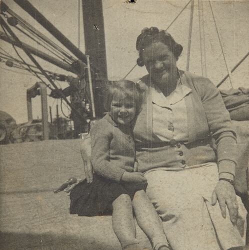 Girl and woman smile, sitting on a ship deck. Ship rigging behind them.