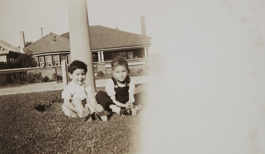 Two young children sit on grass. House can be seen behind them.