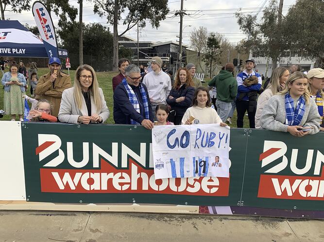 Group of spectators standing behind football oval fence. Girl holds handmade banner over fence with green Bunnings signage. Banner is white with blue letters.