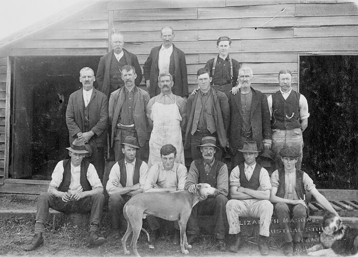 Shearers and shed hands pose in front of a shearing shed. Dog in front centre of group. Dog at right also.