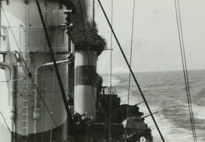 Military ship with smoke coming from one of the ship's funnels, ocean in the right background.
