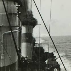 Military ship with smoke coming from one of the ship's funnels, ocean in the right background.