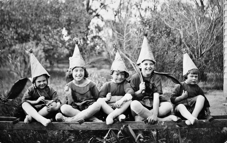 Five girls dressed as pixies sit cross-legged wearing peaked hats. Trees behind.