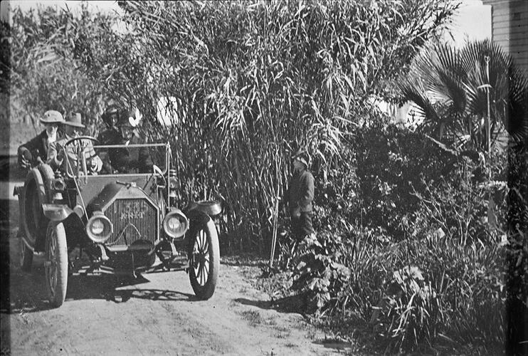 Three people seated in a motor vehicle on a driveway through a garden.