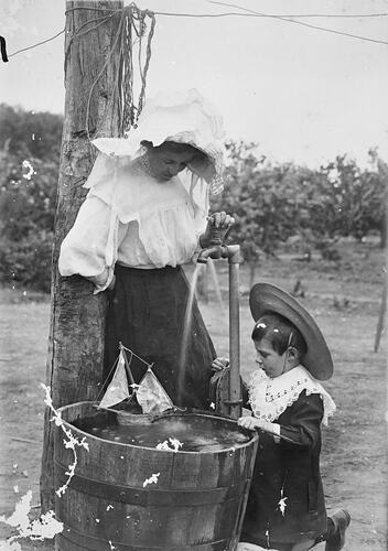 Small boy, kneeling, and a woman, standing, playing with a toy boat in a wash tub. The tub is below two taps in the garden.