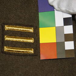 Detail, black cloth square badge on khaki fabric. Features column of three gold horizontal bars.