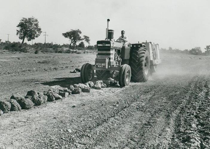 Man driving a tractor towing a roller with large concrete weights on board over a row of rocks.