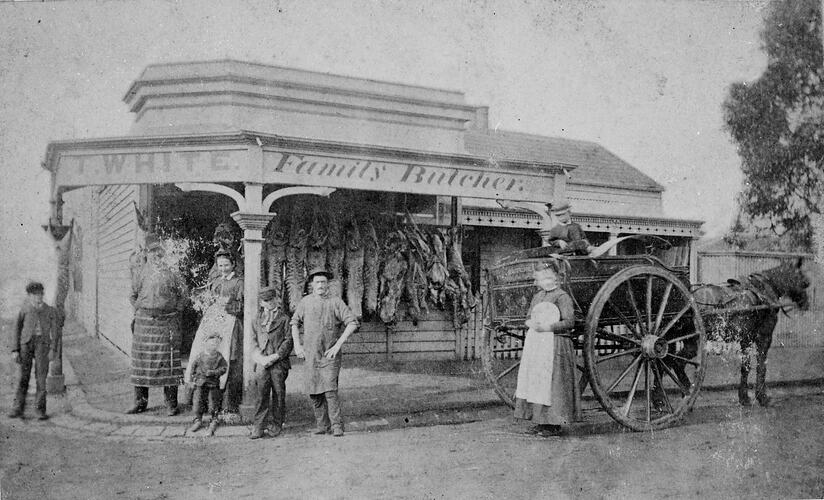 Butcher's family outside their corner shop. Carcases hang from the front wall and delivery cart at right.