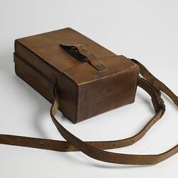 Rectangular leather case with buckle shoulder strap and leather fastening strap. Text stamped on top.