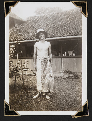 Man standing in sarong and hat with building behind.