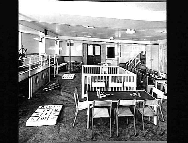 Ship interior. Children's playroom. Small table and chairs at front. Playpen behind. Number game on floor.