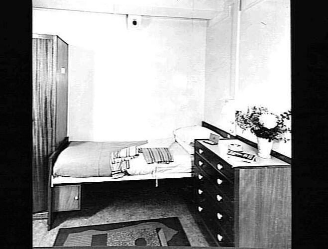 Ship interior. Single bed against wall. Chest of drawers at right.