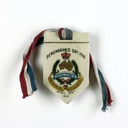 Badge in shield shape with a red, white and blue ribbon knotted through slot at the top, text and image on bad