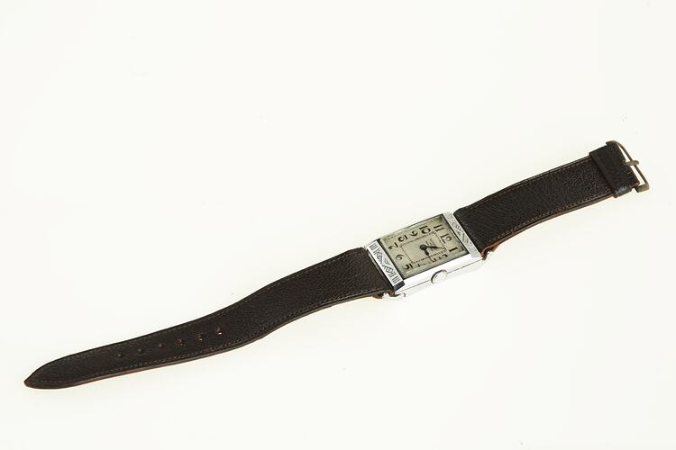 Watch with rectangular chrome case. White metal face has black arabic numerals. Black leather band.