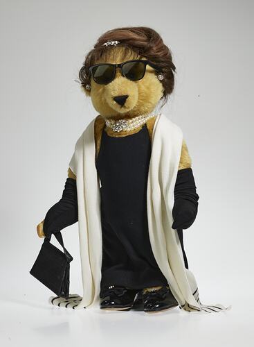 Light brown bear wears black hat, sunglasses, dress, full-length gloves, bag, shoes, white pearls and scarf.