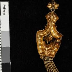 Back of gold pendant earring. Comprised of four connected sections with floral and leaf motifs. Gold fringe hangs from the bottom.
