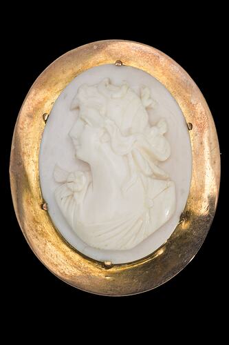 Oval white cameo of female bust facing left. She wears a draped garment and has ribbons in her hair. Plain gold setting.