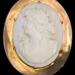 Oval white cameo of female bust facing left. She wears a draped garment and has ribbons in her hair. Plain gold setting.