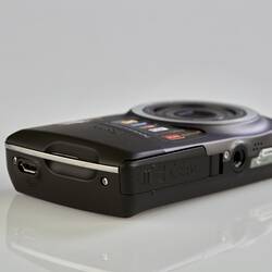 Black and grey camera laying flat, lens up. Base has screw in hole for tripod. Metal trim on sides and lens.